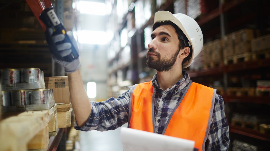 A warehouse operative in PPE using a barcode scanner among the racking of a logistics warehouse to take stock and identify dead stock.