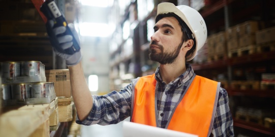 A warehouse operative in PPE using a barcode scanner among the racking of a logistics warehouse to take stock and identify dead stock.