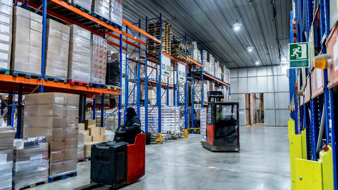 An electric manual pallet truck of the Equipment-as-a-Service type is parked in a large industrial warehouse next to racking filled with goods.