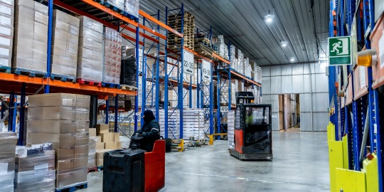 An electric manual pallet truck of the Equipment-as-a-Service type is parked in a large industrial warehouse next to racking filled with goods.