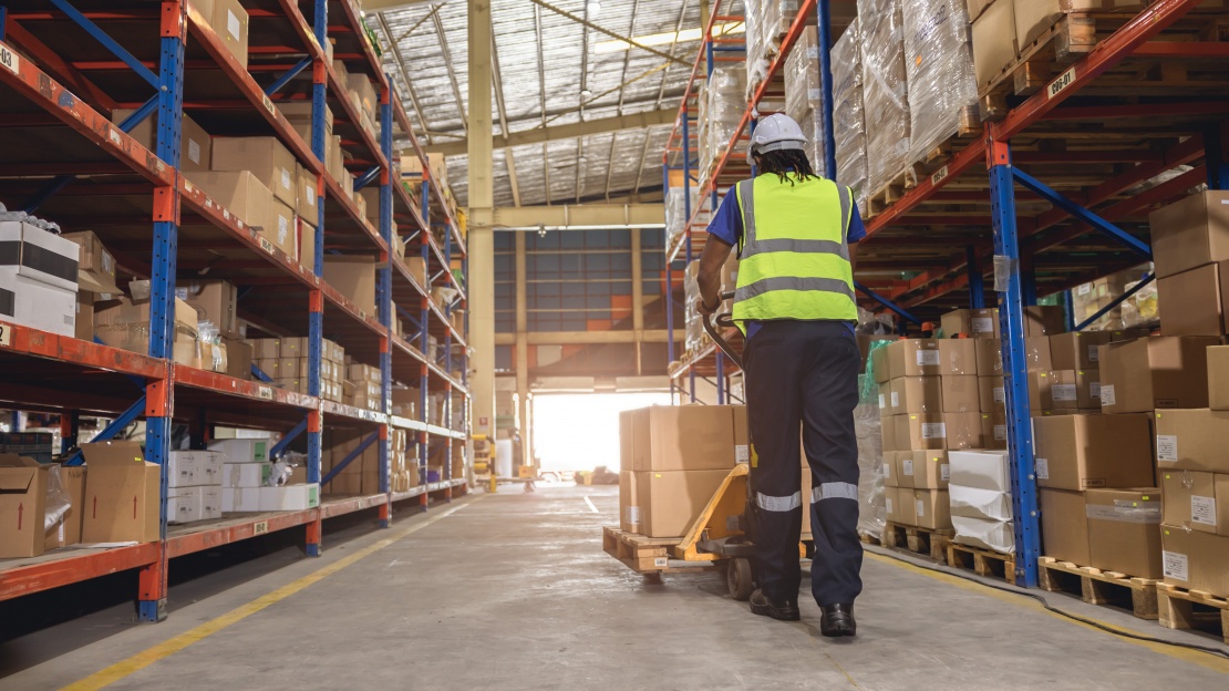 Employé d'entrepôt vu de dos, portant un casque et un gilet de sécurité, tirant un transpalette manuel dans une allée bordée de rayonnages pour optimiser le lead time.