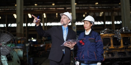 Un manager en costume pointant du doigt dans une usine industrielle tout en discutant de la vérification des green claims de l'entreprise avec une technicienne portant un casque.