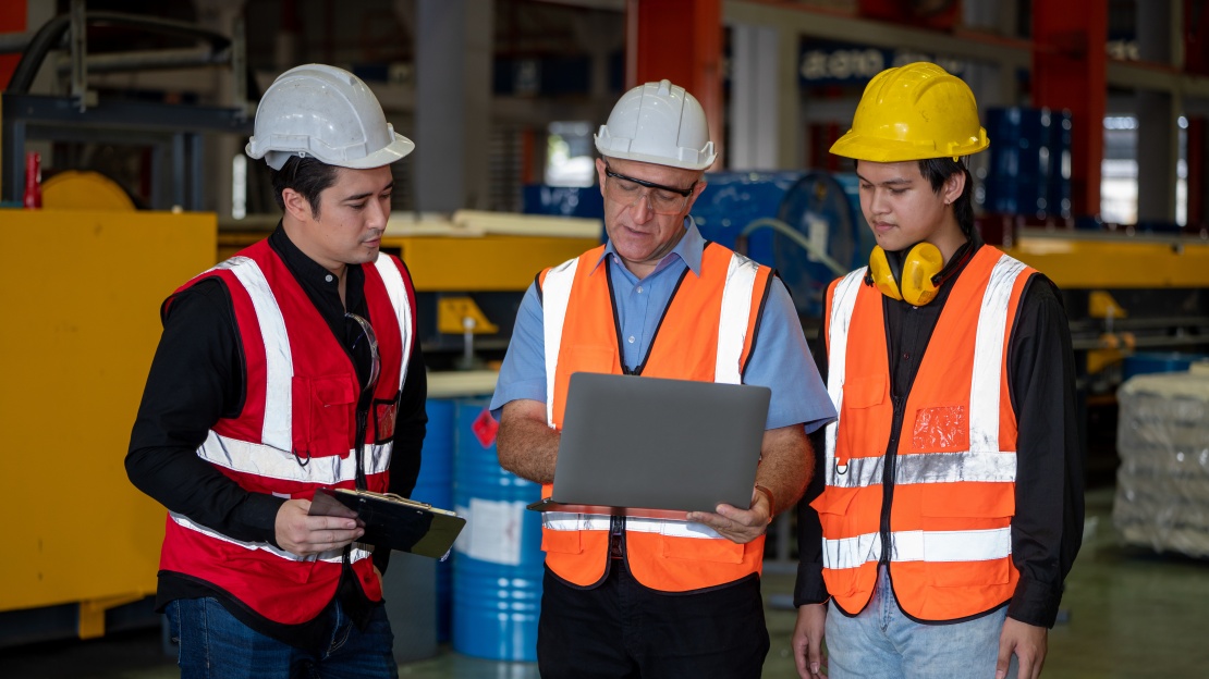 A team of three professionals wearing high-visibility vests gather around a laptop in a factory to schedule preventive maintenance operations.