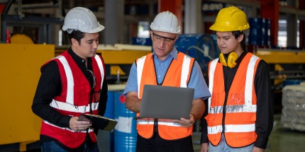 A team of three professionals wearing high-visibility vests gather around a laptop in a factory to schedule preventive maintenance operations.