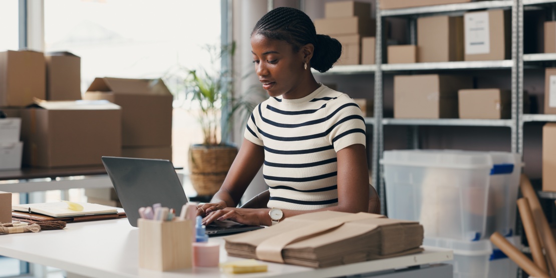 Une spécialiste en logistique travaille sur son ordinateur portable à son bureau entouré de cartons pour planifier une stratégie d'approvisionnement proactif.